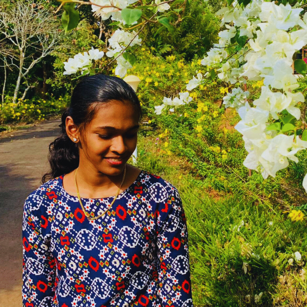 A young woman standing outdoors near blooming white flowers on a sunny day, smiling softly while looking down, used to represent a digital marketer in Calicut in a natural setting.